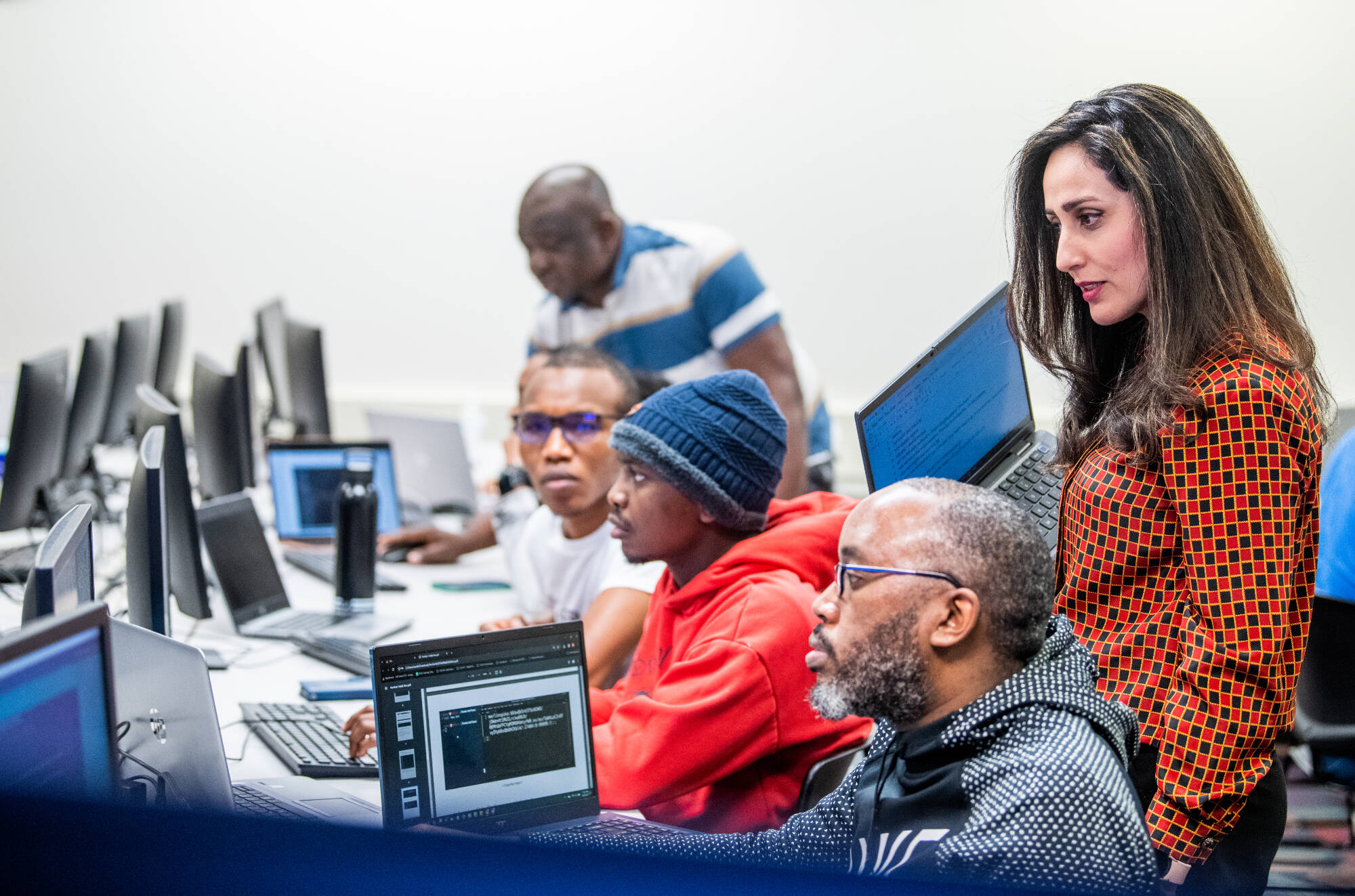 Classroom with computers and students with the teacher looking over their shoulder.